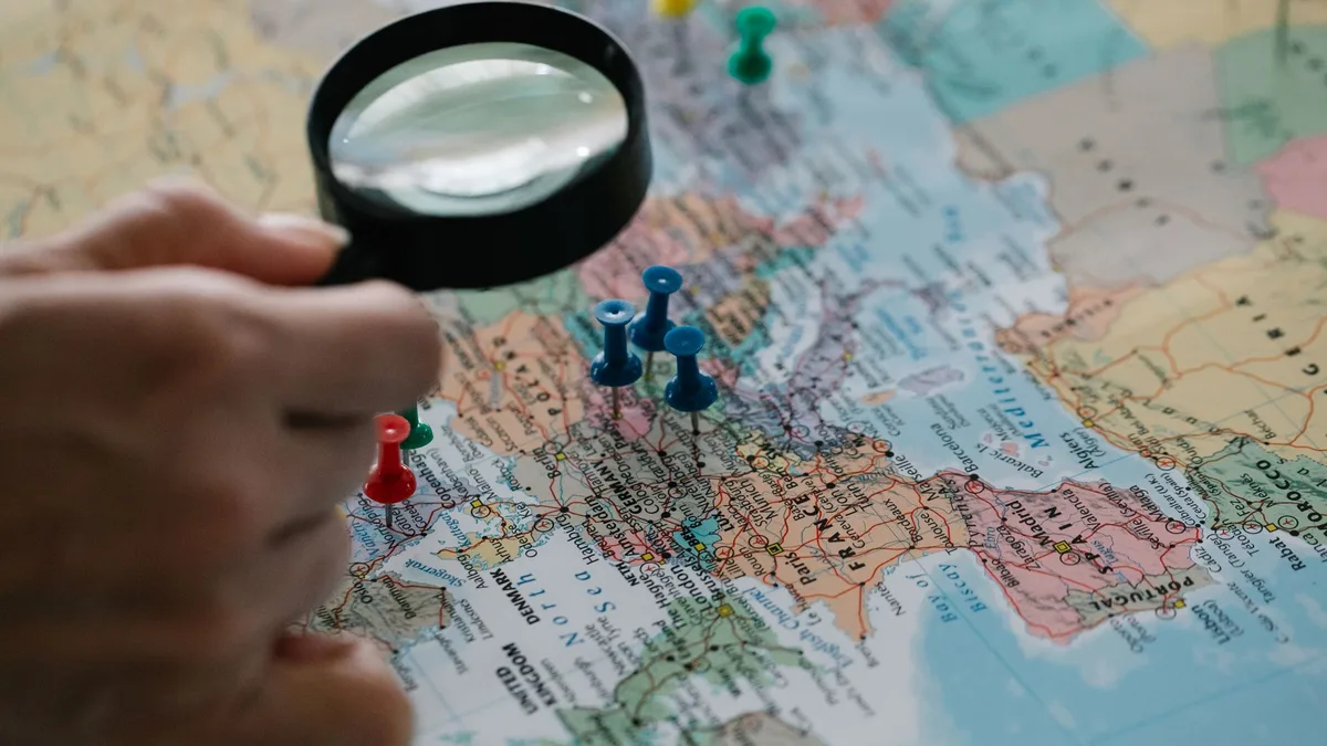 Dłoń z lupą nad mapą świata z kolorowymi pinezkami wbitymi w region Europy i Turcji