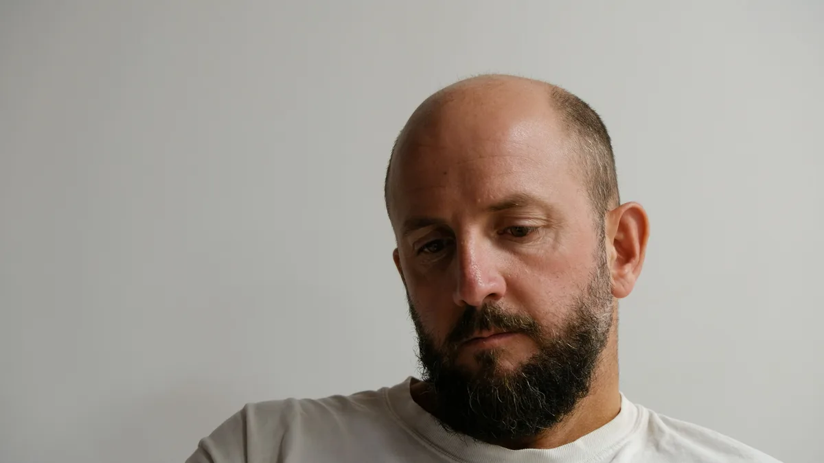 Łysiejący mężczyzna z brodą, w białej koszulce, patrzący w dół z zatroskaniem
